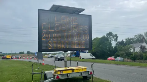A large temporary electric sign saying 'lane closures 20:00 to 06:00 start here' standing right by a road on a very grey day
