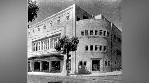 Project Gaumont Black and White shot of the building. It looks very sturdy and lots of brickwork. There is a grand entrance with steps leading up to the doors. There is a small-ish tree outside the building which is on the corner of a road.