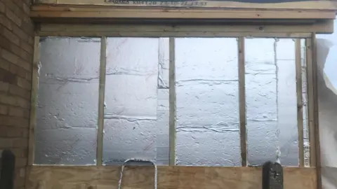 Submitted A wall has had its outer covering removed, revealing wooden battens and silver insulation panels beneath. In front of the exposed section, there is a grey concrete fence post.