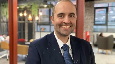 A man wearing a blue jacket with blue tie and white shirt smiling to the camera. He has short brown hair and it is receding.