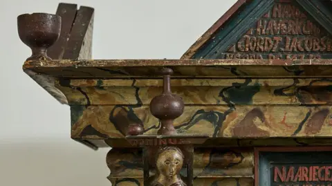 Sworders The corner of an ornately carved cupboard, with lettering and what looks like a wooden wine glass carved out of the wood.