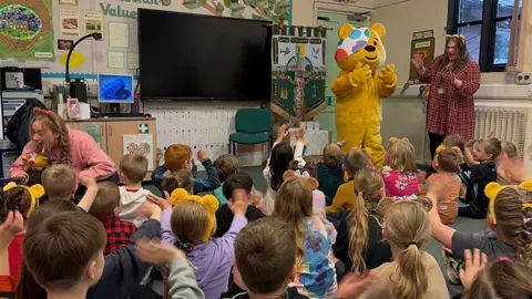 Schoolchildren sitting on the floor with their backs to the camera in a classroom waving at Pudsey Bear who is standing next to a teacher at the front of the class