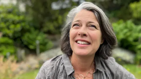 A lady with shoulder length grey hair looking towards the camera. She's wearing a grey blouse and a necklace with metal hoops.