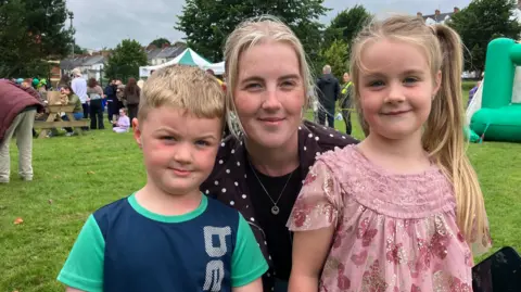 A mum and her two children smile at the camera. They are standing on a patch of grass. Behind them stands a crowd of people. There is also a bouncy castle. 