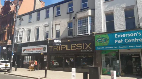 The former Triplesix Studios site, still with the business's shopfront and branding, but with a "To Let" sign tacked to it. Above it are two more floors, with glass windows looking out onto the city centre street. On its right is Emerson's Pet Centre and on its right is Pman Afro Mart. Both businesses have similar spaces above them, and all three are painted white.
