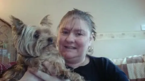 Karen Bassett A woman with tied back dark hair wearing a black top is pictured holding a small Yorkshire Terrier dog. The pair are positioned in a cream-coloured room. 