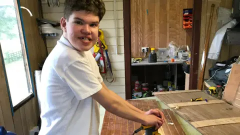 Family photo of Fraser in his dad's workshop holding a tool used to sand down his sailing boat.