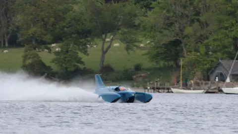 Bluebird K7 "planes" on Coniston Water. The blue craft is throwing up a large spray behind it as it glides on top of the lake's surface. On the far shore, sheep are grazing on an incline.