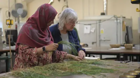 Two women are inside lab sorting through grass cuttings on a table in front of them while consulting an open book
