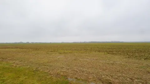 Universal Destinations and Experiences An empty green field at Kempston Hardwick. The sky is grey. Trees can be seen on the horizon.