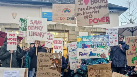 Jess Bailey A crowd of people holding up signs against any cuts to the library service outside Ottery library