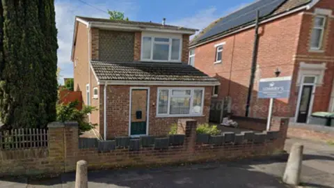 Google A two-storey, brick, detached house, much shorter and less wide than its neighbours, with a "for sale" sign outside