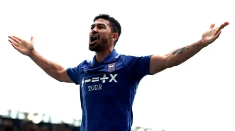 PA Media A football player celebrates scoring a goal. He wears a blue football shirt and has his arms raised out from his body as he shouts in celebration.