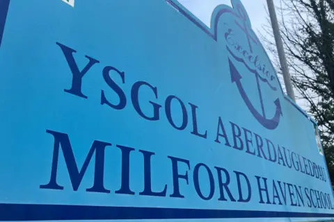 A close up of the school sign which is blue and reads: Ysgol Aberdaugleddau / Milford Haven School with an anchor logo above which features the word Excelsior.