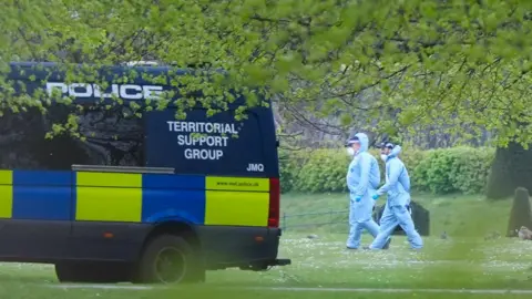 Two men in hazmat suits walk across the grass, shrouded by the branches of a tree. They walk towards a police van parked nearby.