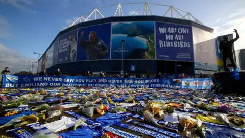 Getty Images Cardiff City Stadium with blue, Bluebirds branding on the outside of it. In front of the stadium are hundreds of bunches of flowers, messages and blue Cardiff City scarves and shorts strewn on the ground. A long blue banner wrapped round the gate in front of the stadium reads: "We never saw you play and never saw you score, but Emiliano our beautiful Bluebird, we will love you forever more." A metal statue of a man with one arm raised in the air is on a plinth to the right of the shot. The sky is blue, it is a clear day. 