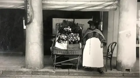 A black woman in old-fashioned clothing and a large hat, stood next to a wicker cart displaying flowers. She is between two pillars in the black and white image.