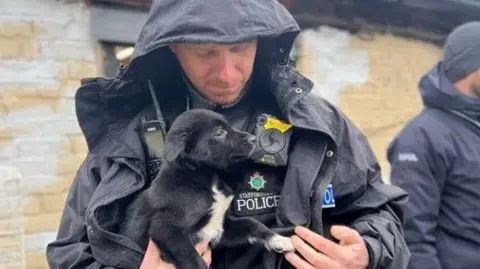 A police officer in uniform holds a black and white puppy. The rescued puppy is reaching out with its paw to the man's hand.