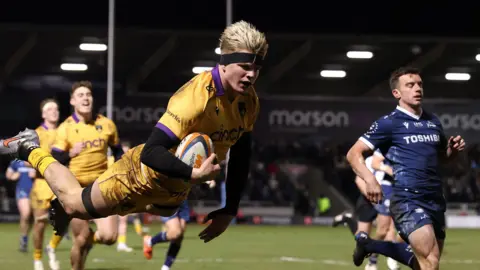 Henry Pollock, wearing a black headband, leaps in the air with the ball under his arm, to score Northampton's final try as Sale fly-half George Ford watches on