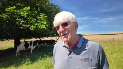 James is wearing a grey polo shirt with dark sunglasses. He is standing in front of a tree with a dozen cows behind him in the shade of the tree.