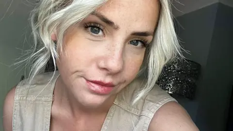 Megan Lynch A selfie of a woman in her 20s with blond wavy hair and red lipstick, who is wearing a beige dress. 