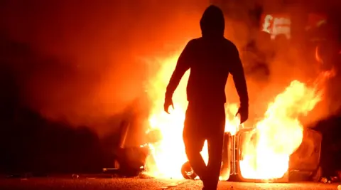 A masked rioter stands in front of what appears to be a wheely bin set alight during riots