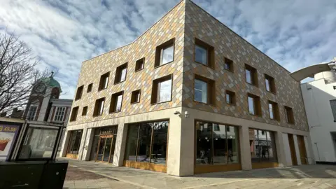 Aimee Dexter/BBC A large three-storey building is in the middle. The first floor has large windows and an entrance on the left. The top two floors have a diamond pattern on the outside. 