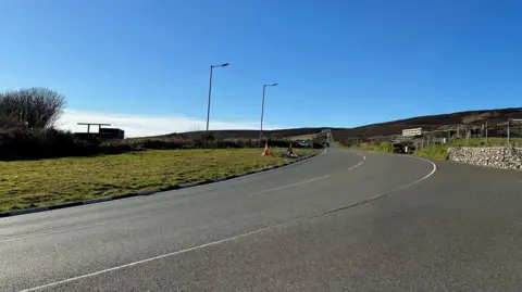 BBC The road sweeping round at the Creg ny Baa wit4 the road heading off towards Kate's Cottage. There is blue sky above.