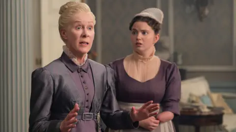 Netflix A screenshot from Bridgerton shows a woman with blonde hair in a bun looking shocked next a woman with brown hair looking worried. The woman with brown hair has a limb difference as she does not have a forearm or hand. They are both wearing purple dresses. 