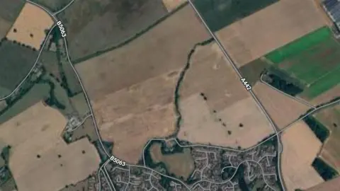 An aerial google maps view of two large brown fields with a link of greenery between them. To the house are hundreds of houses. Bordering it are two main roads - the A442 on the right and the B5063 on the left
