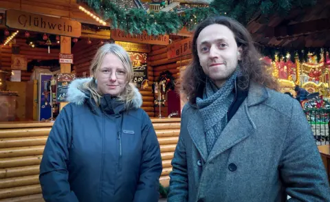 A woman with blonde hair in a black coat and a man with brown hair in a grey coat and grey scarf stand in front of a wooden hut at a Christmas market that sells alcohol, adorned with fake Christmas tree sprigs, fairylights, signs saying "Gluhwein"