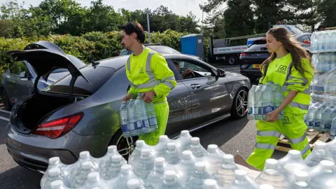 Getty Images A man and a woman in yellow high-vis clothing load cases of bottled water into an open car boot. There are pallets of bottles beside them.