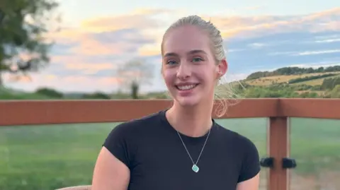 Young woman with blonde hair, wearing a black short-sleeved shirt and green pendant necklace. She is sitting in countryside with fields and trees in the background.