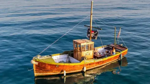 Darren Loud Aerial view of the wooden boat in the sea. It has a flat stern, a central wheelhouse and a flagpole bearing a jolly roger pirate flag.