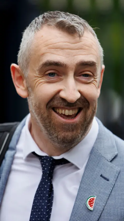 Daithi McKay, a man in a grey suit and blue tie, smiling broadly