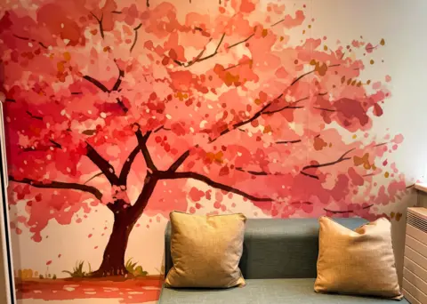 WISHH A brightly-coloured modern tree design on a wall next to a seating area. It has reddish leaves shooting off from the branches.