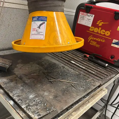 An industrial bench used for welding. Above it is a yellow ventilation system, and to the right is a red box with a black handle.