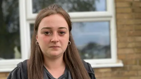 Abigail Swinn frowns as she stands outside her home in front of a window. She has long brown hair and is wearing a dark, hooded top and silver small-hooped earrings. 