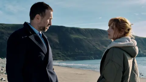 Sky Rafe Spall, left, and Kelly Reilly, right, are facing each other with a beach and hills in the background. Spall wears a blue shirt and navy blue coat, while Reilly wears a green coat with a fleeced-lined hood