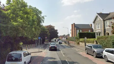 Google A wide main road with cars, houses and trees lining each side. Traffic lights, street lamps and road signs can be seen in the background.