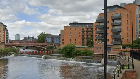 A view of the Rive Aire going through Leeds city centre. On the right are tower blocks and in the distance is a bridge