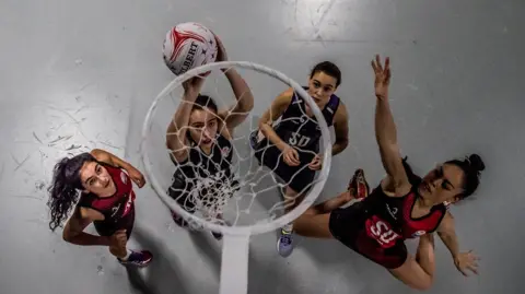 Shot from above of a netball hoop with four female players beneath. The woman second from left is about to put a white ball into the hoop. Furthest right (Farah Hasan) is sticking out her arm to try to block the ball. The others are looking at the hoop. There are all wearing dark blue and red netball vests and shorts. 