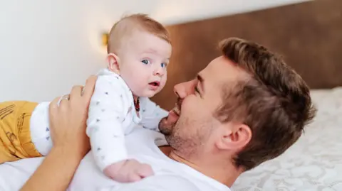 Getty Images A man is lying on a bed holding a baby. The baby is looking at the camera, while the dad is looking at the baby.