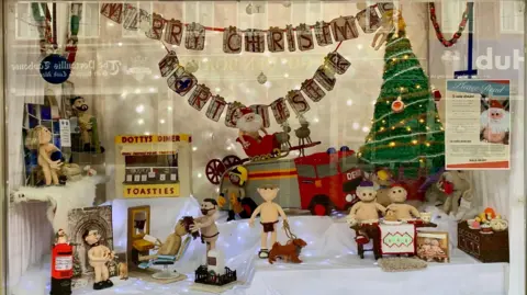 the nude knitted characters on display in a window with Christmas decorations hanging all around