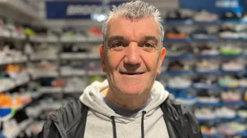 Sports shop owner, Richard Knox, wearing a grey hooded jumper, a black jacket and smiling in front of a room full of trainers