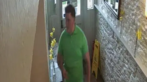 A white man is wearing a green polo shirt and is walking in a narrow hallway. Behind him is a door and a wet floor sign.