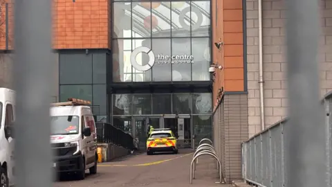 BBC An entrance to The Centre Livingston at Almondvale Shopping Centre, showing a police vehicle and a white van parked near the glass-fronted doorway.