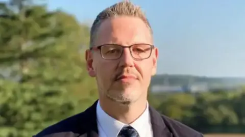 A man with short hair and glasses in a black suit and white tie with blurred shapes of trees behind him