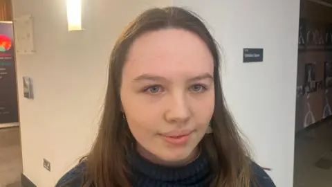 A close up picture of a girl looking directly in to the camera. She is wearing a navy roll neck jumper. She has brown hair and blue eyes. There is a white wall in the background. 