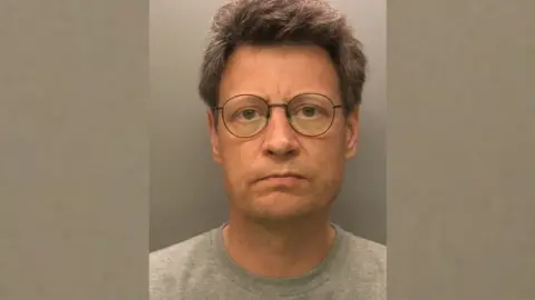 A mug shot of Daniel Holden. He has short grey hair and wears thin-rimmed black glasses and a grey t-shirt. 